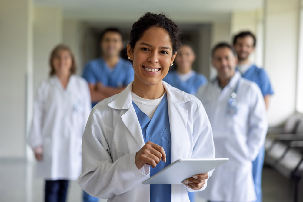 Gestão em Saúde: 3 opções de pós-graduação. Descrição: gestora de uma área de saúde na frente usando jaleco branco com equipe atrás com cinco pessoas ao fundo ou de jaleco ou com roupa cirurgica. 